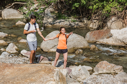 Active young hikers, man and woman walking over a mountain stream flowing among large beautiful rocks. Concept of nature exploring.