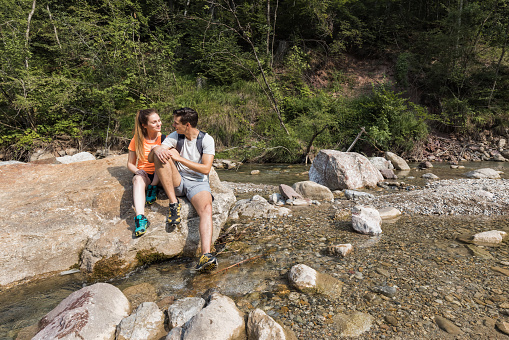 Young couple taking a break from hiking, sitting on the mountain river bank and watching beautiful nature