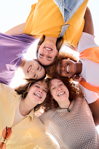 Vertical Group of only girls young cheerful multiracial friends looking at camera hugging happy in community. Diverse females in circle looking camera. Low angle of view empowered women students