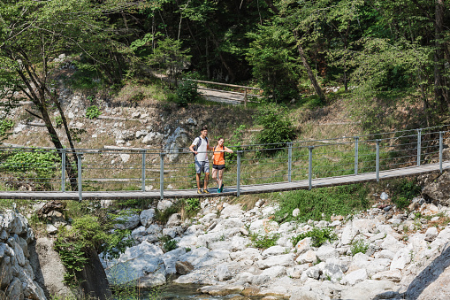 Young heterosexual hiking couple crossing the suspension bridge over a mountain river during summer hiking tour