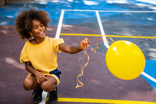 A young girl is playing on a basketball court with her yellow balloon.