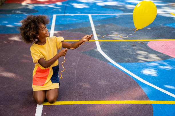 An African-American girl is outside, on a basketball court, trying to fly her yellow balloon. She is smiling and having fun.