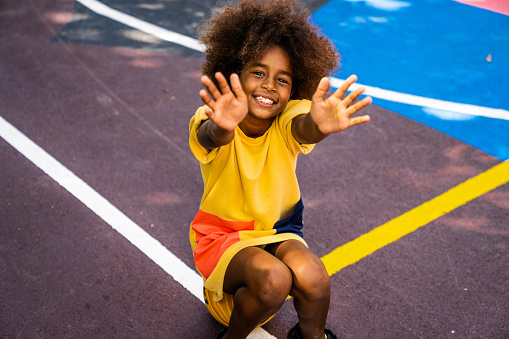 An African-American girl is reaching out her hands while sitting on her ball at the basketball court.