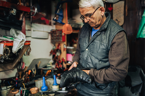 A senior cobbler is examining footwear he is about to repair while standing at his workshop. An old craftsman is standing at his shoemaker workshop and examining boots for reparation. Craftsmanship.