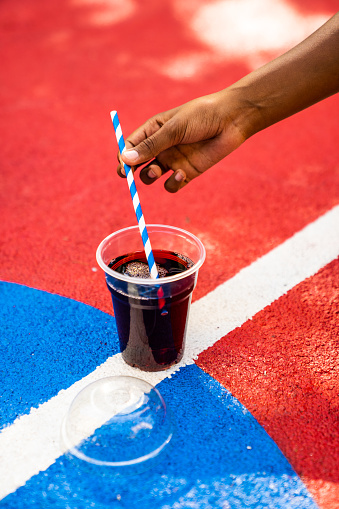 A cup of juice to go is on the basketball court. There's a cup's lid on the ground next to it. A young black girl is mixing the juice with the straw.