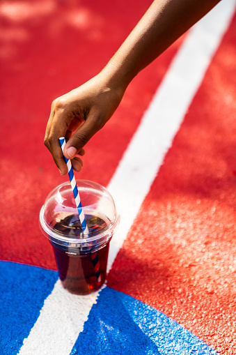 A cup of juice to go with a straw is on the basketball court and a black girl is taking it to have a refreshment on a hot summer day.
