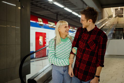 Beautiful couple at railway station or a subway waiting for the train. Traveling together. Love, friendship and public transport. Woman holding man for his hand and they are looking at each other.