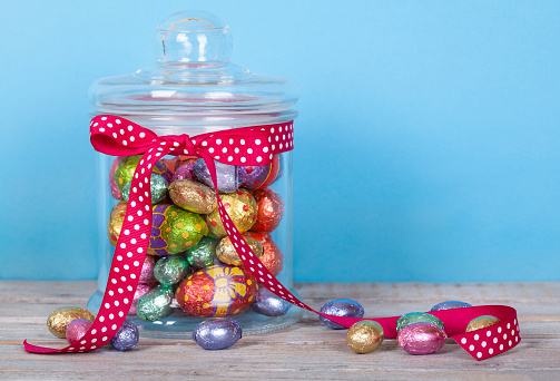 Colorful Chocolate Easter Eggs in Glass Jar on white rustic wooden table.