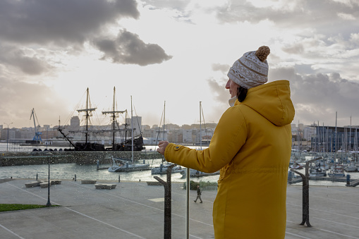 a woman in a vibrant yellow raincoat, her back turned to the camera, as she gazes out towards the harbor. The scene is set against a backdrop of a dramatic sky, where the sun peeks through the clouds, casting a golden glow that reflects off the water's surface. A historic tall ship, its sails furled, stands majestically at the dock, serving as a reminder of the bygone era of sailing. The harbor is alive with the movement of smaller modern boats, juxtaposed with the stillness of the grand ship. This moment is a blend of contemplation and admiration, as the woman seems to be lost in thought, appreciating the harmony between the nautical past and present.