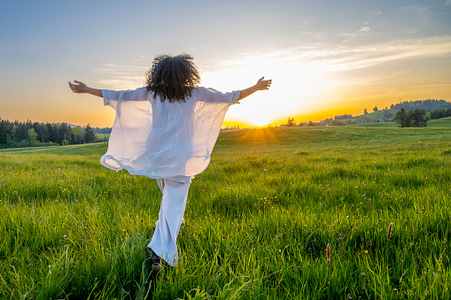 Latin woman spreading hands and running in summer field at sunset, wide shot back view