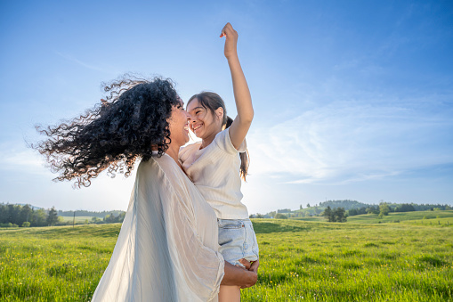 Happy smiling mother holding and hugging daughter in summer field at sunset, medium shot