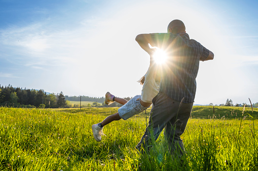 Father holding daughter and spinning around in field at sunset, wide shot