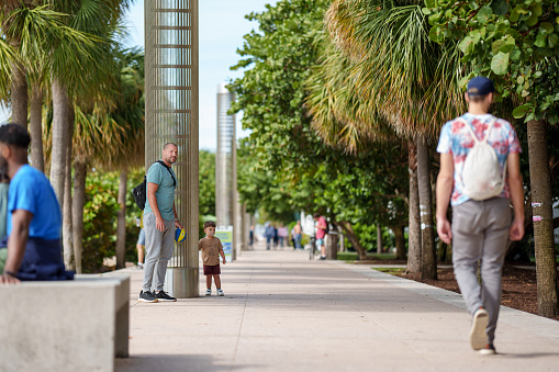 A Caucasian man stands with his Puerto Rican three year old son on a busy walking path in a tropical city on a beautiful day in Florida.