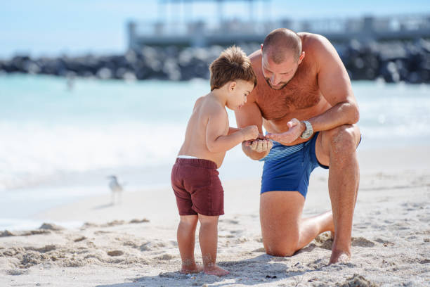A Puerto Rican three year old boy stands next to his Caucasian father as they look at a shell they found together while spending the day at the beach in Florida.