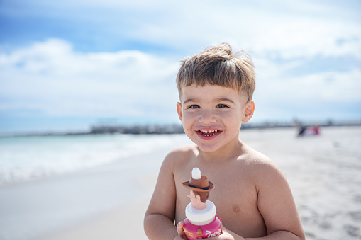 An adorable Puerto Rican three year old boy wearing swimming trunks stands in the sand, eating a snack and smiling to the camera while enjoying a day at the beach.