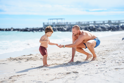 A loving and fit Caucasian man squats next to his toddler son of Puerto Rican descent, playing with each other on the beach on a beautiful day in Florida.