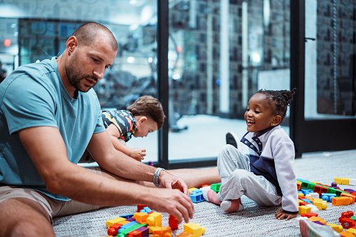 A happy young girl of African American descent smiles at an unrecognizable adult man and a young boy while they play on the floor with toys and blocks together.