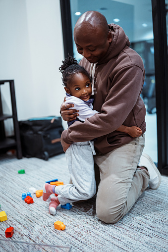 An adorable toddler girl of African American descent smiles while hugging her father as they play with blocks together on the floor inside their home.