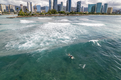 A wide, aerial view of an unrecognizable surfer sitting on their surfboard in the ocean, with the city of Honolulu, Hawaii in the background.