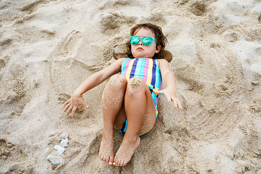 An adorable four year old girl of Eurasian descent wearing sunglasses and a swimsuit lays in the sand on the beach while enjoying a sunny day in Hawaii.