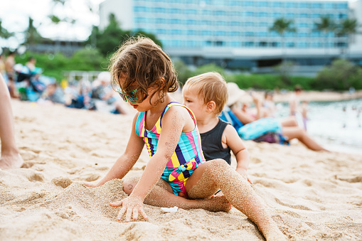 A cute four year old Eurasian girl sits with her young brother in the sand, playing together while enjoying a day on the beach during a family vacation.