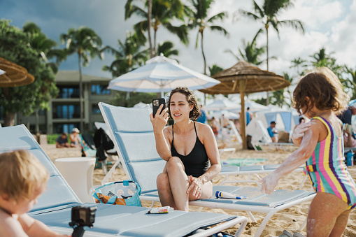 A beautiful woman of Eurasian descent sits on the beach chair, talking on the phone while her toddler daughter applies sunscreen. The two are enjoying a day on the beach in Hawaii.