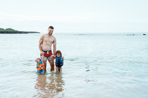 A handsome and muscular man of Caucasian descent holds hands with his Eurasian toddler son and daughter while playing in the shallow ocean in Hawaii.