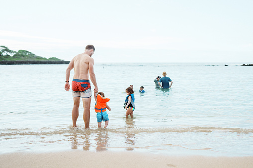 Rear view of a loving Caucasian father holding hands with his one year old, Eurasian son while walking into the shallow ocean on an overcast day in Hawaii.
