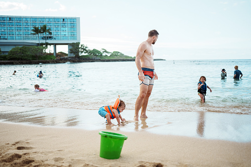 A handsome man of Caucasian descent stands in the sand at the beach, enjoying a vacation in Hawaii with his Eurasian toddler son and daughter.