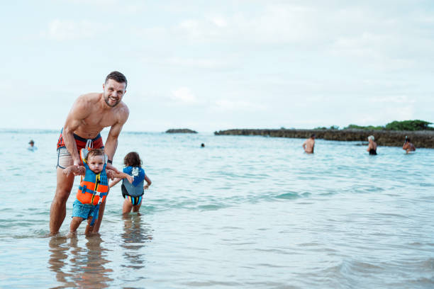 A handsome man of Caucasian descent and his Eurasian one year old son smile to the camera while enjoying a day playing in the ocean during a tropical family vacation in Hawaii.