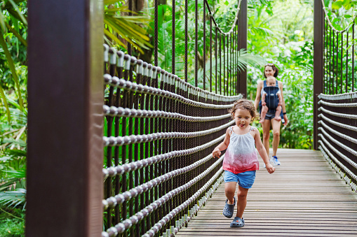 A multiracial, three year old girl runs on a suspension bridge with wonder as she walks through a garden with her mother and brother.