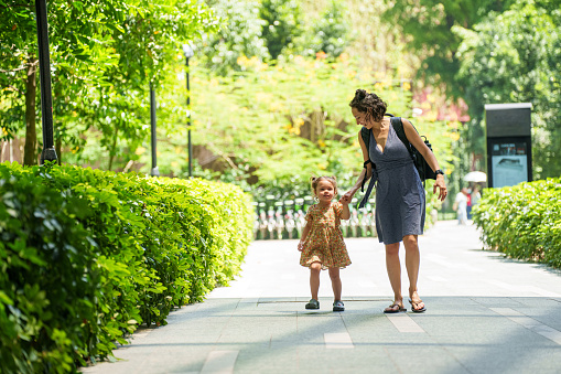 An adorable multiracial four year old girl holds hands with her mother while taking a walk together through a lush park in Asia.