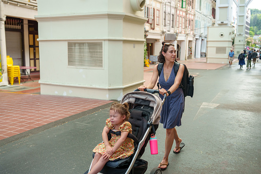 A beautiful woman of Eurasian descent pushes a stroller, holding her toddler daughter through a city in Asia during a family vacation.