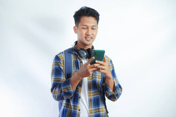 istock Smiling happy young Asian man student in casual clothes backpack using mobile phone, chatting online and reading news isolated on yellow background. high school university college concept 2038994911