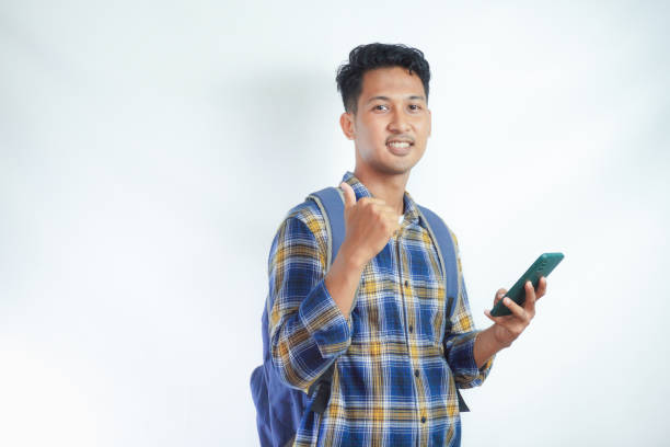 istock Cheerful young Asian man student in casual clothes backpack standing while holding mobile cell phone and showing thumb up isolated on white background. high school university college concept 2039004491