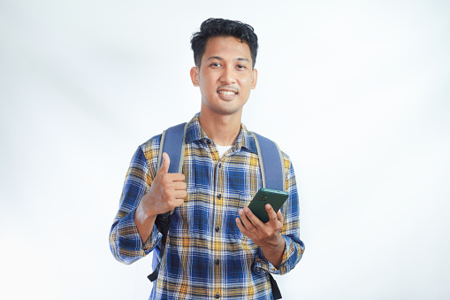 Cheerful young Asian man student in casual clothes backpack standing while holding mobile cell phone and showing thumb up isolated on white background. high school university college concept