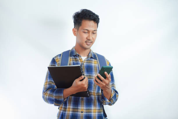 istock Smiling happy young Asian man student in casual clothes backpack holding book using mobile phone, chatting online and reading news isolated on yellow background. high school university college concept 2039009254