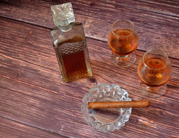 cognac, strong alcohol in a crystal decanter and two glasses on a dark wooden table, next to it is a cuban cigar in a glass ashtray. - whisky liqueur glass alcohol bottle imagens e fotografias de stock