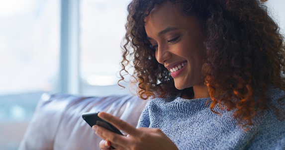 Relax, happy and phone for social media with a woman reading on a sofa in the living room of her home. Mobile, contact and smile with a young person chilling in her apartment while typing a message