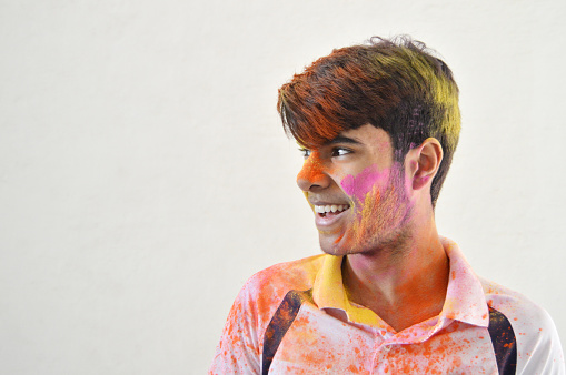 Horizontal portrait of one happy smiling young 16 year old boy's side view with smile on his face painted messy with Holi powder colours over gray rough wall background with copy space