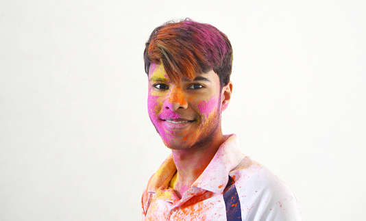Horizontal portrait of one happy smiling young 16 year old Indian Asian boy's portrait headshot with smile on his face painted messy with Holi powder colours over gray rough wall background with copy space