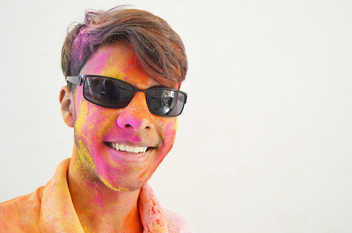 Horizontal front view portrait of one stylish cool happy smiling young 16 year old boy wearing black sunglasses or goggles with smile on his face showing teeth, painted messy with Holi festival powder colours over rough grey colored background