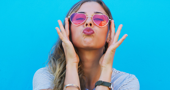 Portrait, heart sunglasses and kiss with a woman on a blue background for love or romance on valentines day. Summer, fashion and pout with a confident young gen z model in trendy eyewear for style