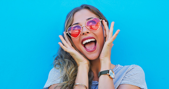 Fashion, heart sunglasses and a woman on a blue background while shouting with energy in excitement. Summer, smile and beauty with a happy young gen z model in trendy eyewear for style freedom