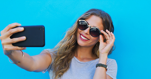 Woman, selfie and sunglasses with smile, wall and fashion for web blog, profile picture or happy by blue background. Gen z influencer girl, photography and memory for post, update or social media app