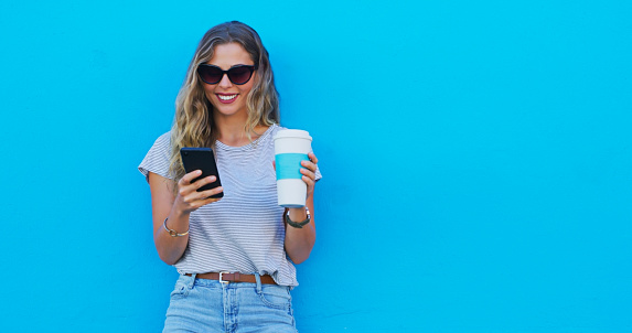 Woman, phone and coffee with space by wall, sunglasses and texting for communication on mobile network. Girl, smartphone and smile with latte, mockup and thinking on social media by blue background