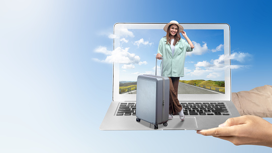 A human hand holding a laptop with an Asian girl in a hat and a suitcase with a hill view and blue sky on the laptop screen. Traveling concept