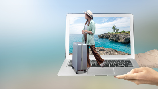 A human hand holding a laptop with an Asian girl in a hat and a suitcase with a sea cliff and an ocean view on the laptop screen. Traveling concept