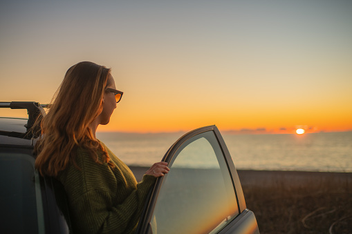 Mid aged woman enjoying sunset on the beach near her car, travel and tourism concept.