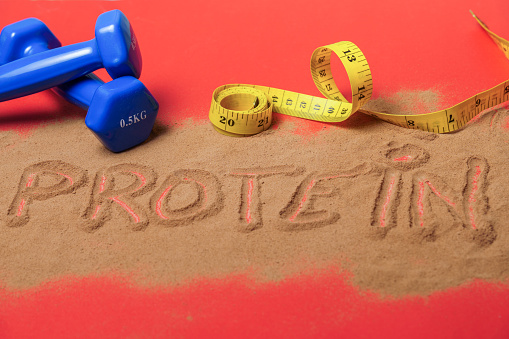 word protein written on protein powder on red background, with dumbbells and measuring tape